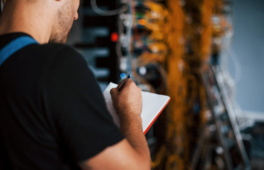 young man in uniform with notepad in hands works with internet equipment and wires in server room